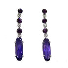 Pair of dark purple crystal drop earrings with three round stones above a long oval stone, silver tone, on white