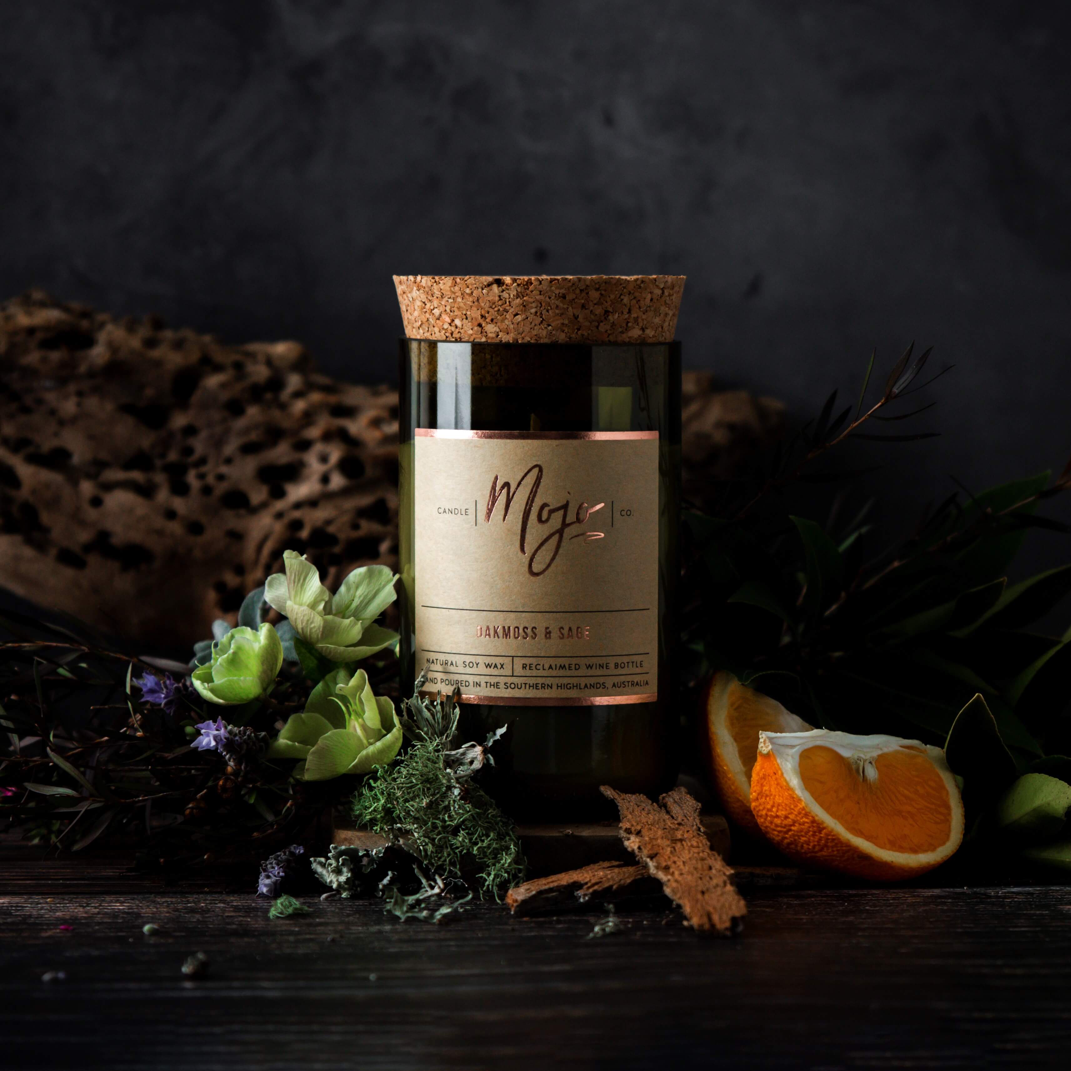 Oakmoss & Sage candle in a reclaimed wine bottle styled with cork bark, herbs and orange on a dark bench