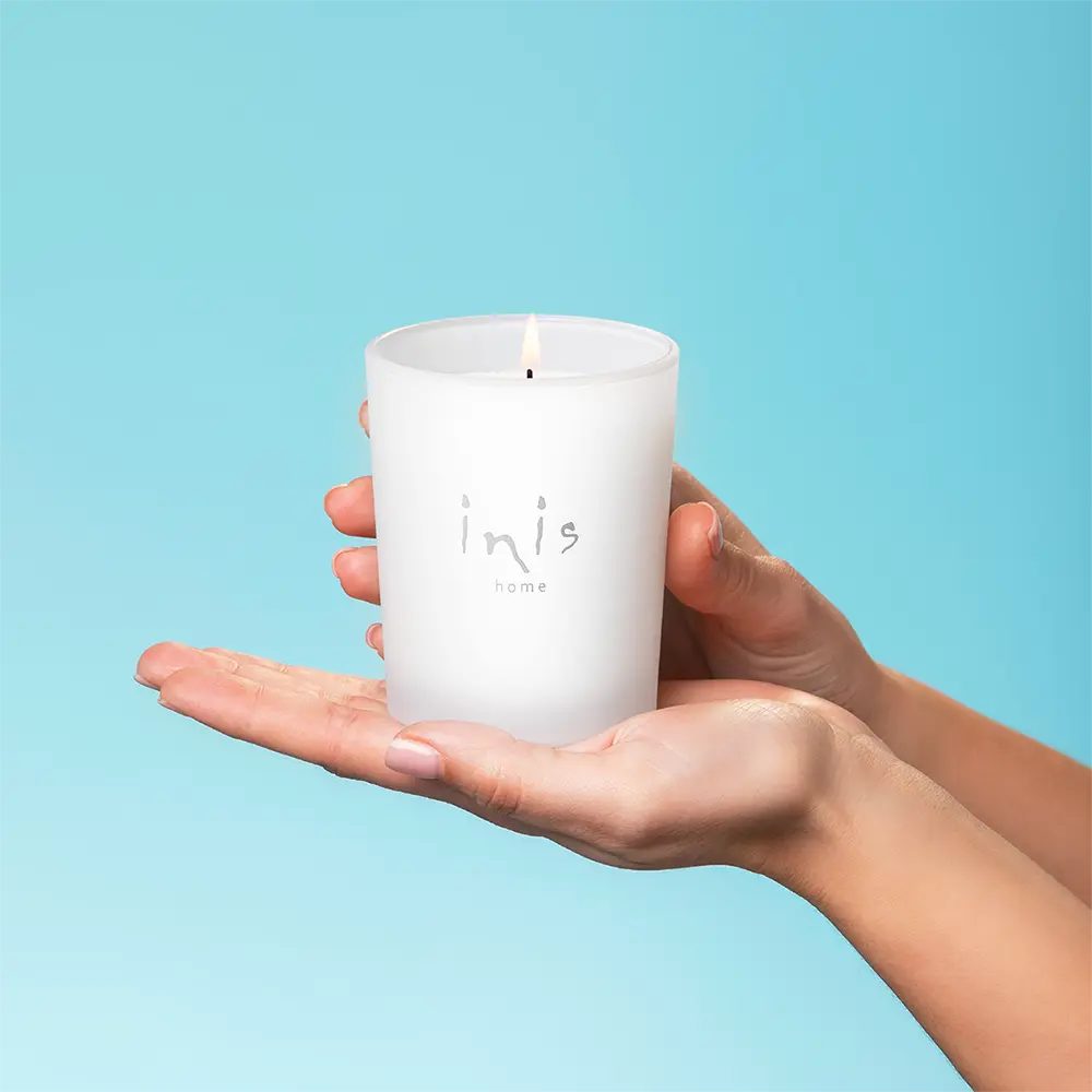 Lit Inis Scented Candle held in two cupped hands against a pale blue background