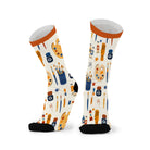 Pair of cream crew socks with a bright orange cuff and black toe, printed with paint palettes, brushes, tubes and ink bottles in ochre, navy and orange.