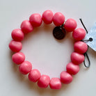 Rock bracelet in Pink: a stretch bracelet of chunky rose pink resin beads with its swing tag and metal charm, laid on a pale surface.
