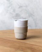 White speckle stoneware travel mug with lid and beige silicone band, on a timber bench