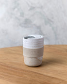 White speckle travel mug with beige band, angled view on timber