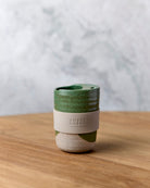 Green dip-glazed stoneware travel mug with beige silicone band, on a timber bench