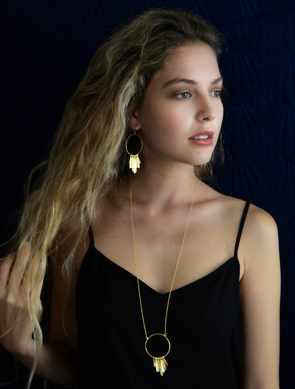 Model in a black camisole wearing the long gold necklace with a fringed circle pendant and a matching earring