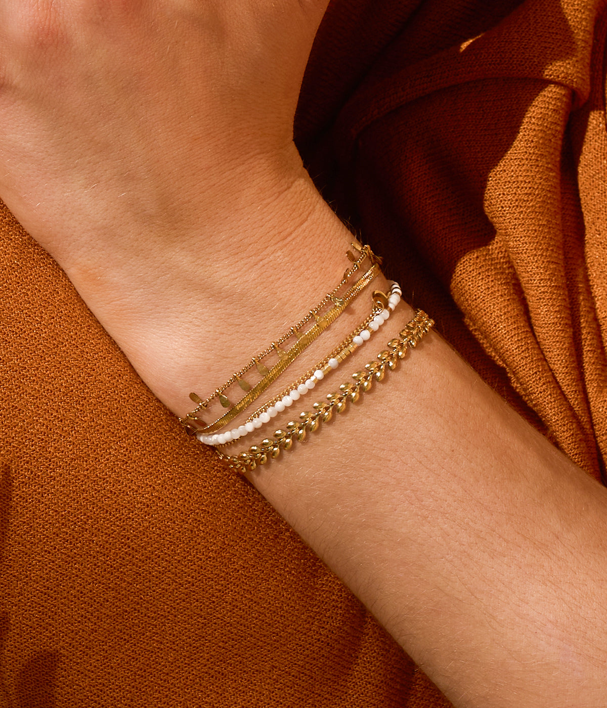 Model's wrist against a rust sleeve wearing the beaded bracelet layered with fine gold chains