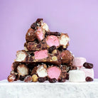 Chunks of Signature Recipe rocky road with pink and white marshmallow, hazelnuts, cranberries and coconut on a purple background