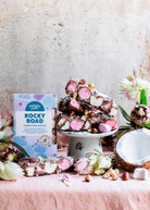 Pebbly Path Signature Recipe rocky road 200 g box beside rocky road piled on a cake stand, with a halved coconut, hazelnuts and marshmallows