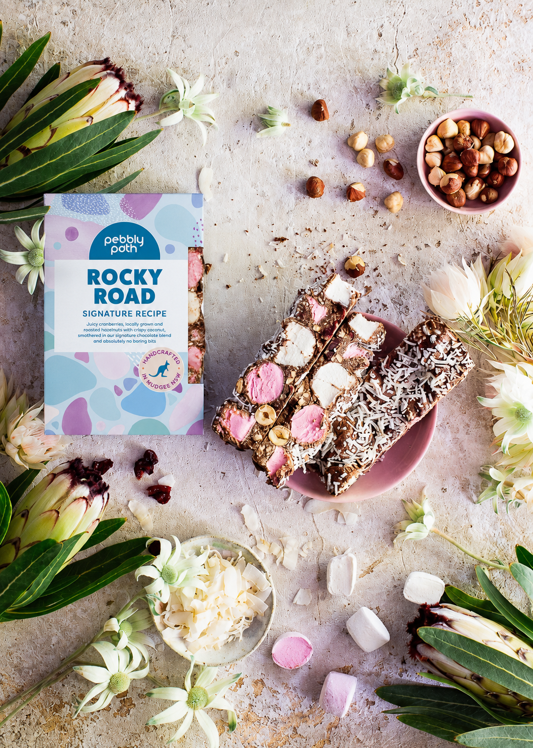 Signature Recipe rocky road 200 g box with cut slices on a pink plate, hazelnuts, coconut flakes, marshmallows and native flowers, seen from above
