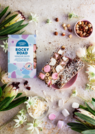 Signature Recipe rocky road 200 g box with cut slices on a pink plate, hazelnuts, coconut flakes, marshmallows and native flowers, seen from above