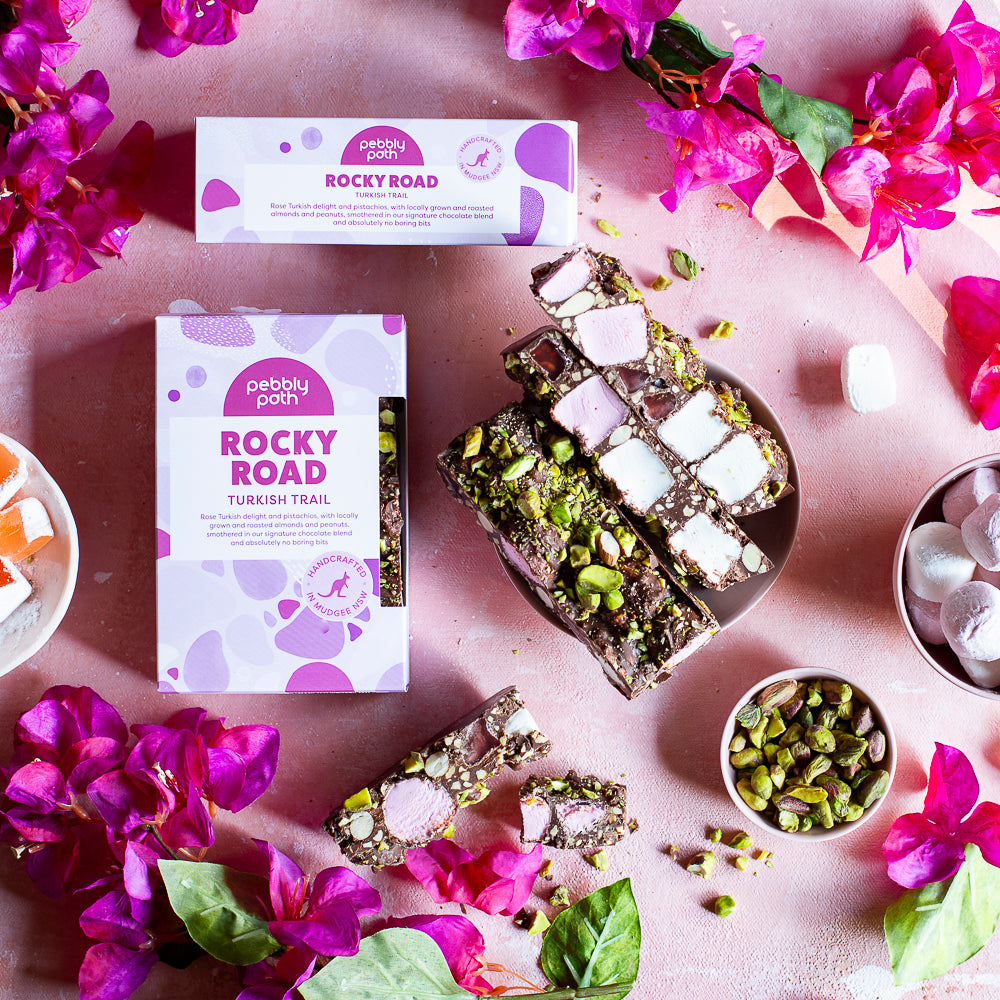 Turkish Trail rocky road slices on a plate with pistachios and marshmallows, beside the 100 g and 200 g boxes and bougainvillea