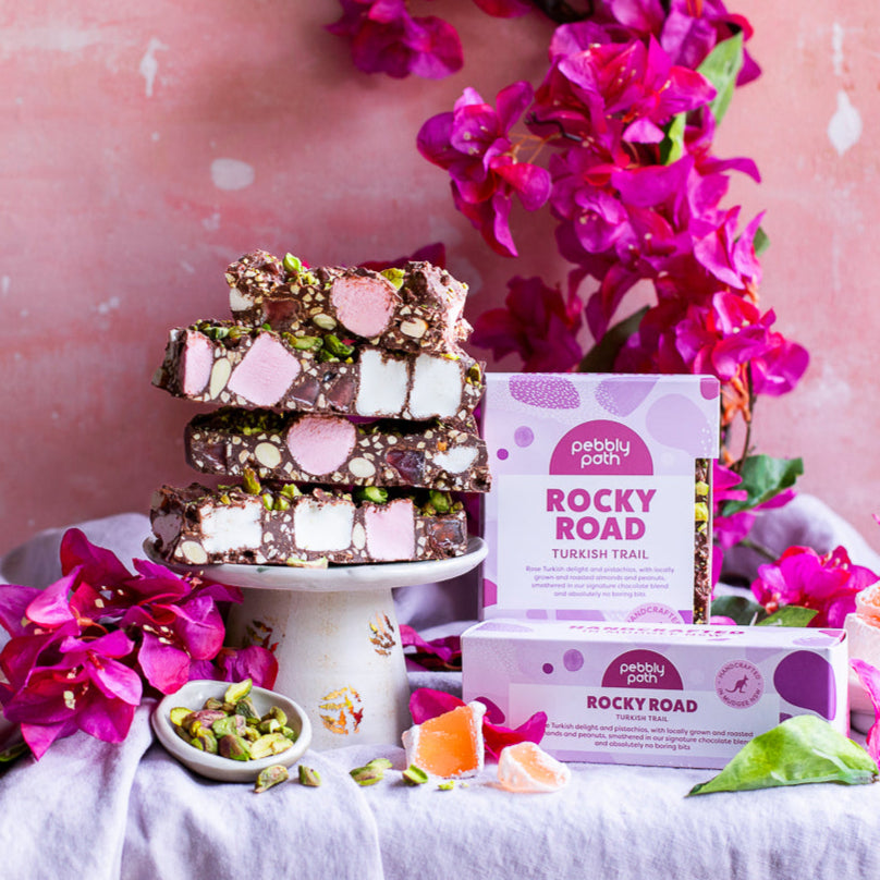 Stacked slices of Turkish Trail rocky road on a cake stand showing pink and white marshmallow and nuts, with the 100 g and 200 g boxes