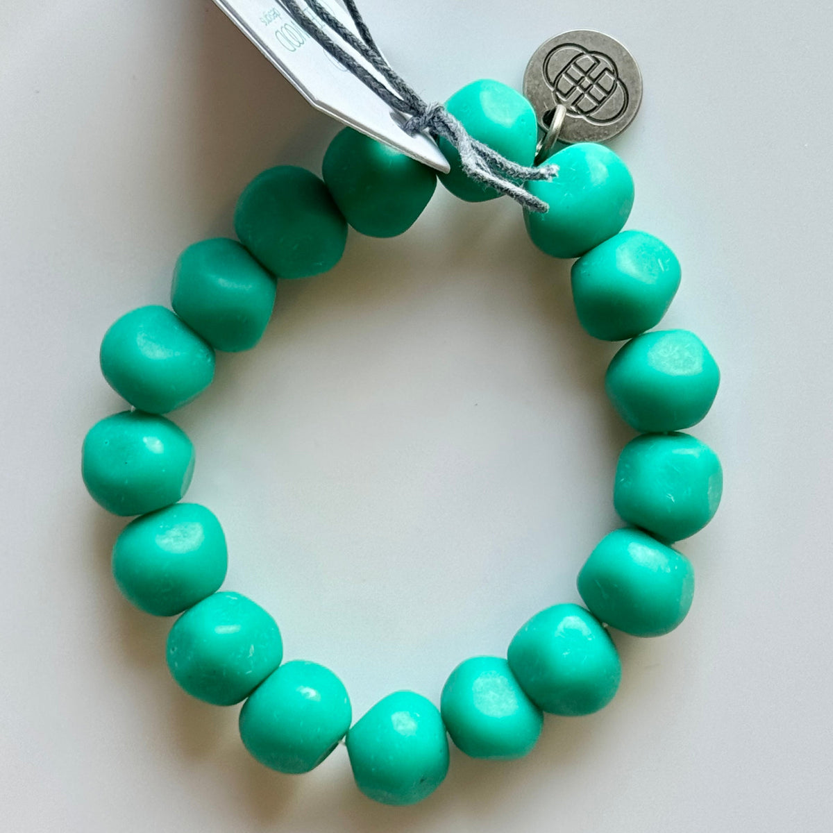 Rock bracelet in Teal: a stretch bracelet of chunky teal green resin beads with its swing tag and metal charm, laid on a pale surface.