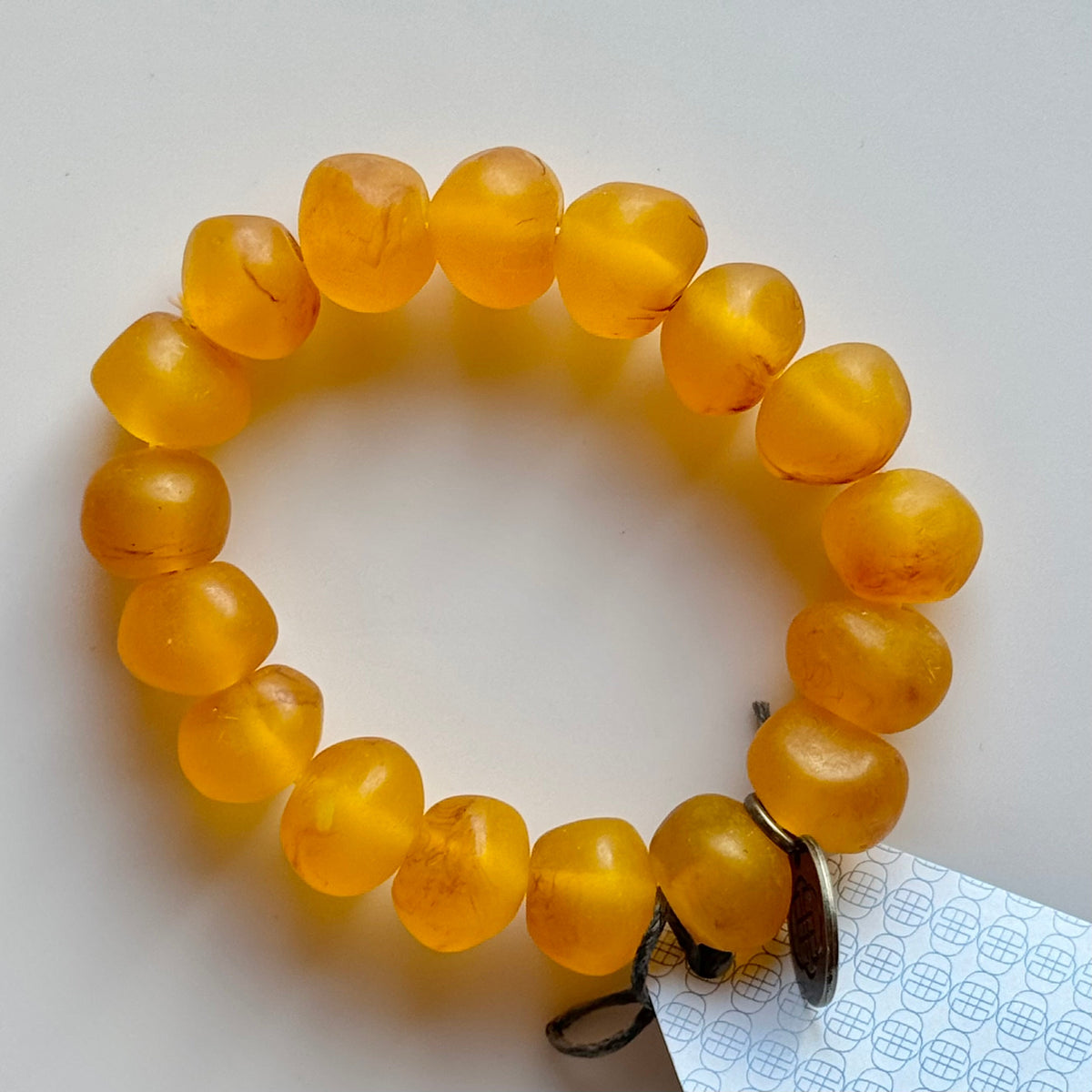 Rock bracelet in Tortoiseshell: a stretch bracelet of chunky translucent amber resin beads with its swing tag and metal charm, laid on a pale surface.