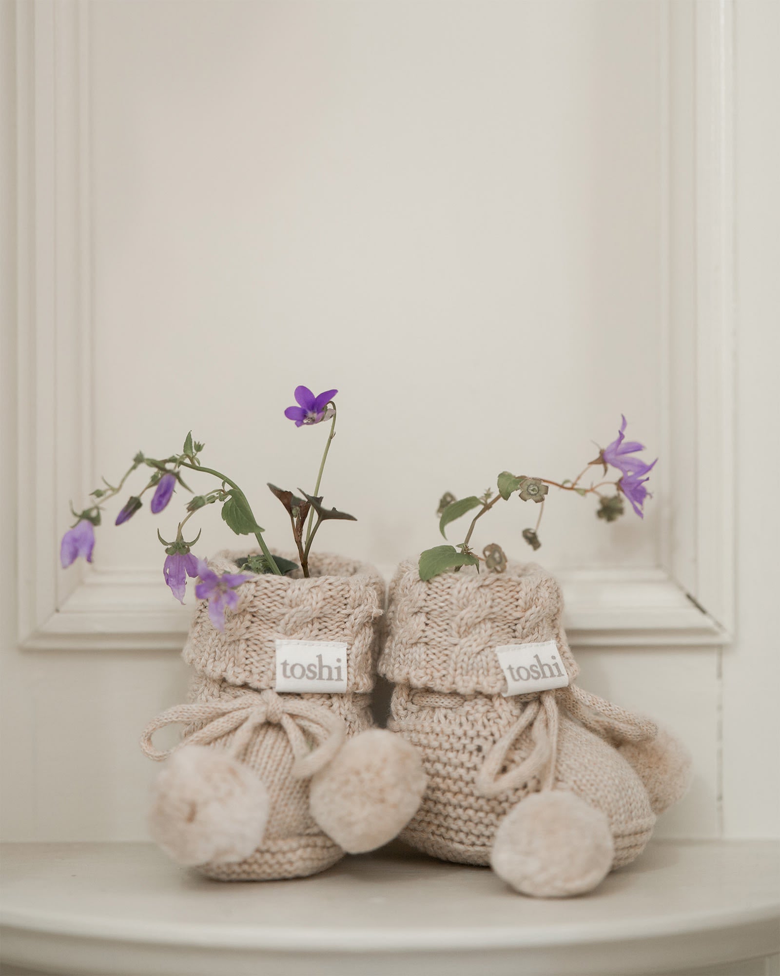 The oatmeal booties on a pale mantel with sprigs of purple violets tucked into the cuffs