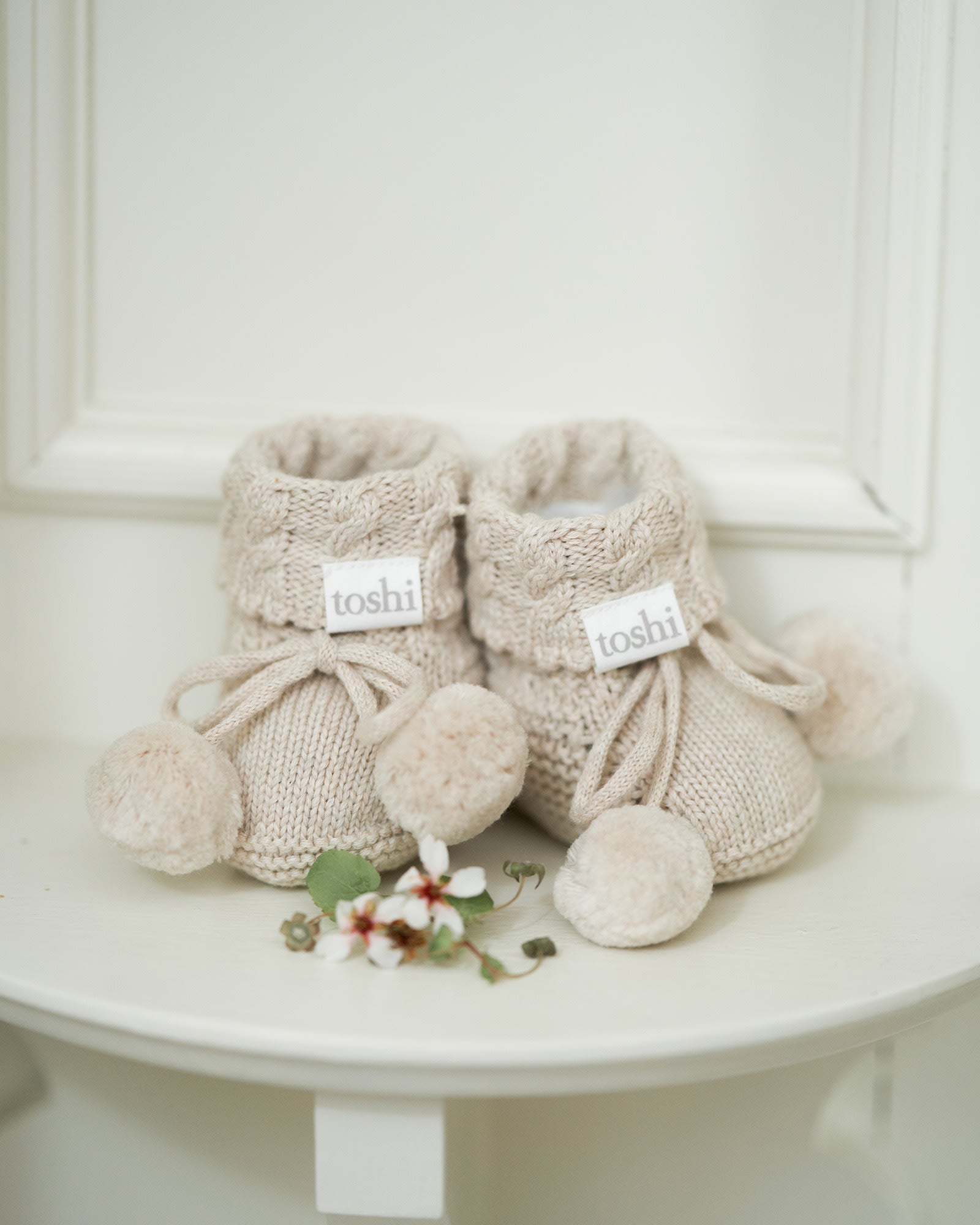 The oatmeal booties on a white side table beside a spray of small white blossom
