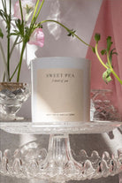 The Sweet Pea candle on a glass cake stand with sweet pea stems behind.