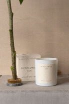 The Sunflower candle and its box on a concrete ledge beside a tall sunflower stem.
