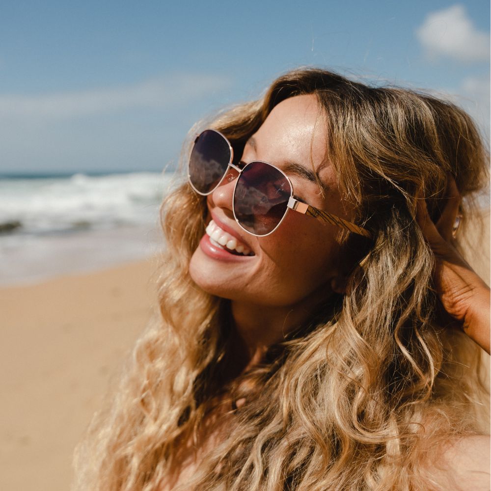 A woman with wavy blonde hair smiling on a beach in the Cleo Bloom sunglasses