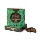 Green sunburst box of dark peppermint bark standing on a second box, with shards of bark flecked with green peppermint beside it