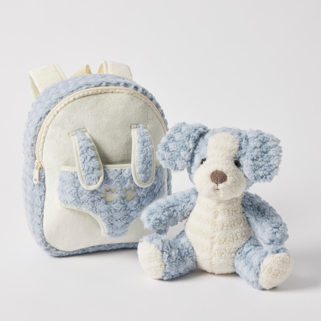 Pale blue curly plush backpack with cream panels and paw prints on the front pocket, the matching blue and cream puppy sitting beside it