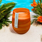 Wave Orange Brazilian Carnival candle on white sand by a pool, with palm fronds and orange blooms