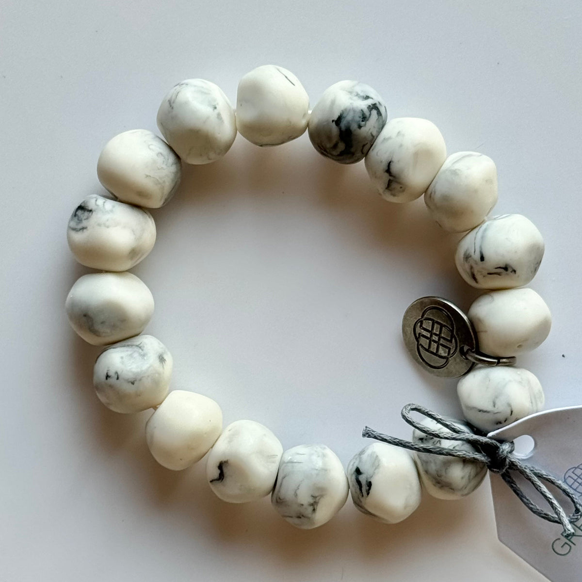 Rock bracelet in White Marble: a stretch bracelet of chunky white and grey marbled resin beads with its swing tag and metal charm, laid on a pale surface.