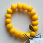 Rock bracelet in Yellow: a stretch bracelet of chunky sunshine yellow resin beads with its swing tag and metal charm, laid on a pale surface.