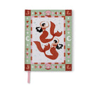 A Dopo journal standing upright, its cover showing two red mermaids inside a sage green border of shells, seahorses and moons, with a pink ribbon marker hanging below