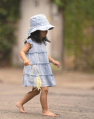 Child in a denim sunhat walking barefoot in the Indiana tiered dress, holding a flower