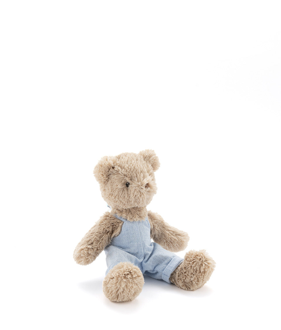 Small light brown teddy bear in pale blue overalls, sitting against white.