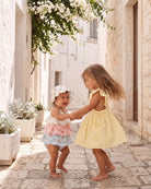 Two toddlers dancing in a sunny lane, one in the lemon Florence pinafore