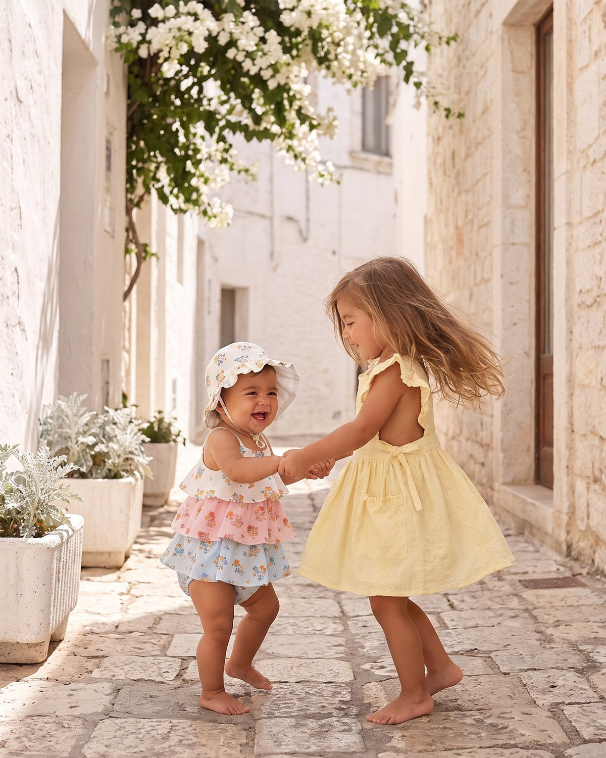 Two toddlers dancing in a sunny lane, one in the lemon Florence pinafore