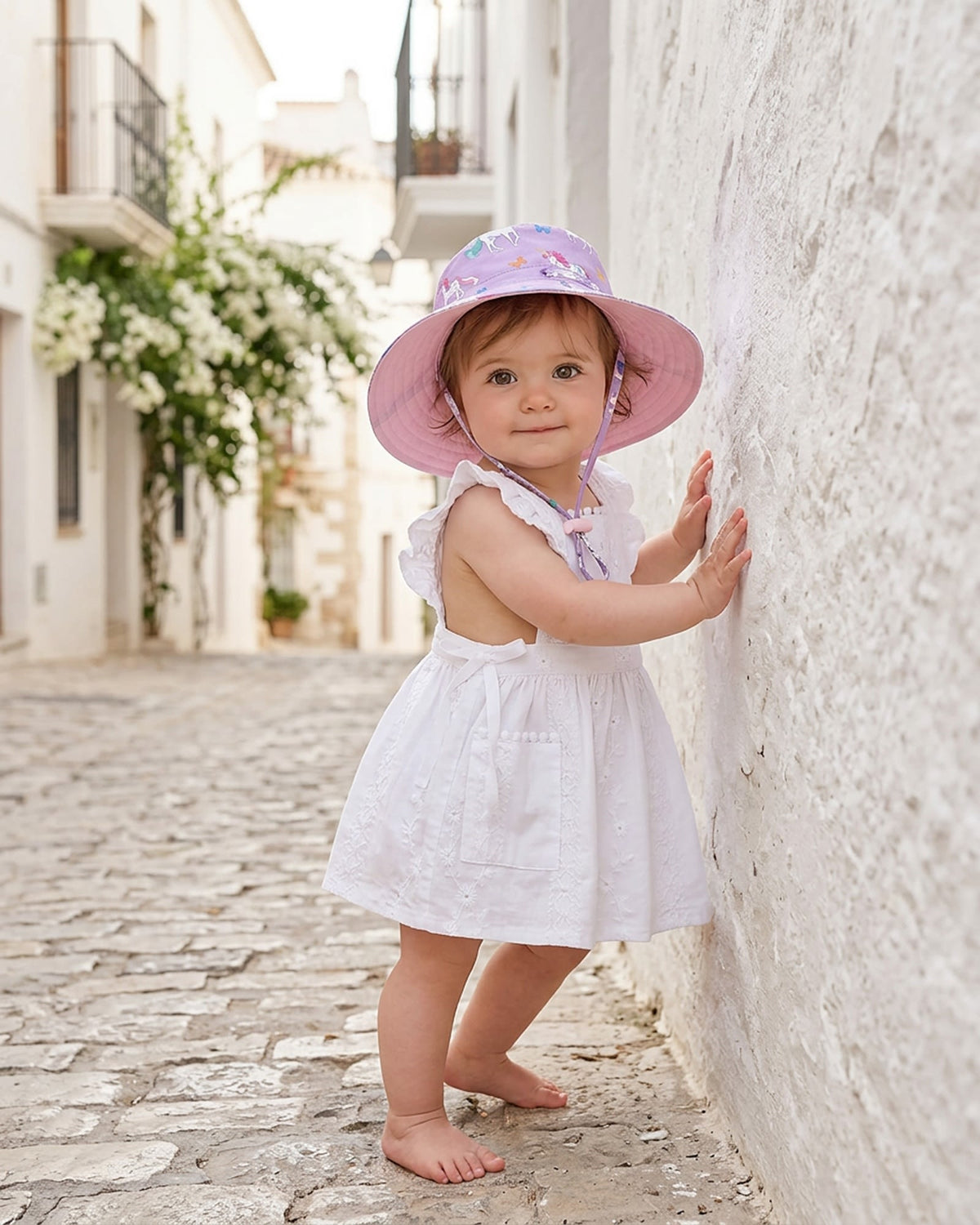 Toddler in the white Florence pinafore and a pastel sunhat, standing against a whitewashed wall