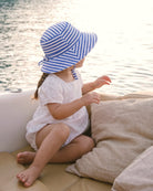 Toddler in the white Florence romper and a blue striped sunhat, sitting on a boat