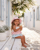 Toddler in the Maddie tiered romper and a pink sunhat, sitting on a white bench with an ice cream