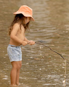 Child standing in shallow water with a stick, wearing the Indiana shorts and an orange sunhat