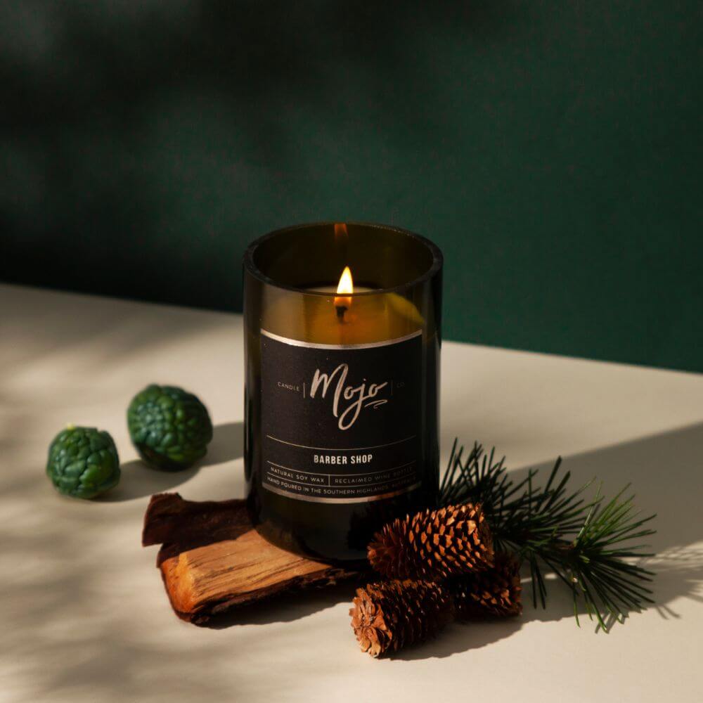 Lit Barber Shop candle in an amber reclaimed wine bottle beside pine cones and timber