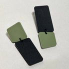 A pair of acrylic earrings on a pale grey surface, each linking an olive green rounded square to a long matte black rectangle, one with the green square on top and the other with the black rectangle on top.
