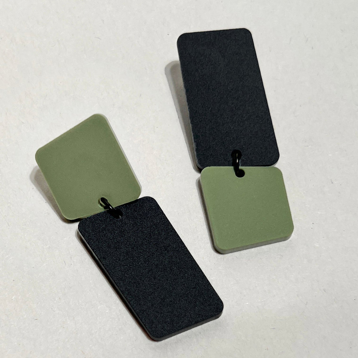 A pair of acrylic earrings on a pale grey surface, each linking an olive green rounded square to a long matte black rectangle, one with the green square on top and the other with the black rectangle on top.