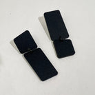 A pair of matte black acrylic earrings on a pale grey surface, each a small rounded square linked to a long rectangle, one hanging square-up and the other rectangle-up.