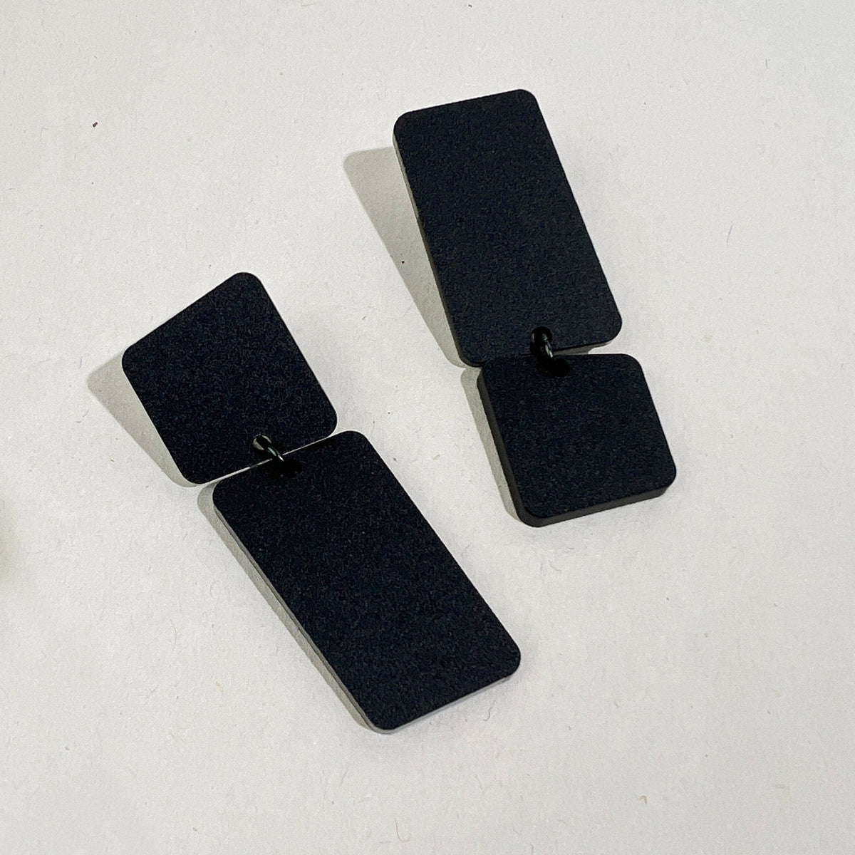 A pair of matte black acrylic earrings on a pale grey surface, each a small rounded square linked to a long rectangle, one hanging square-up and the other rectangle-up.