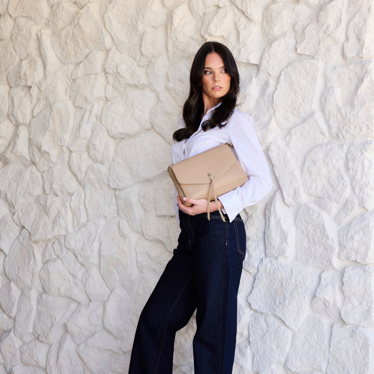 A woman in a white shirt and dark jeans holds the biscuit bag under her arm against a white stone wall.