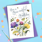 Card hand-lettered Have a Bee-autiful Birthday above painted purple, pink, orange and yellow flowers with bees, on a purple envelope beside a paintbrush and coloured confetti dots