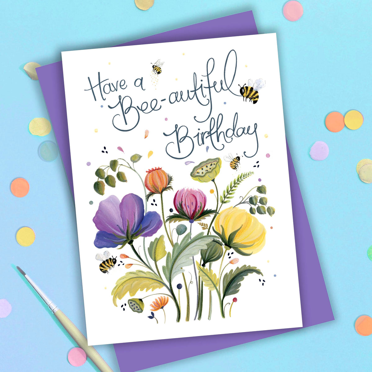 Card hand-lettered Have a Bee-autiful Birthday above painted purple, pink, orange and yellow flowers with bees, on a purple envelope beside a paintbrush and coloured confetti dots