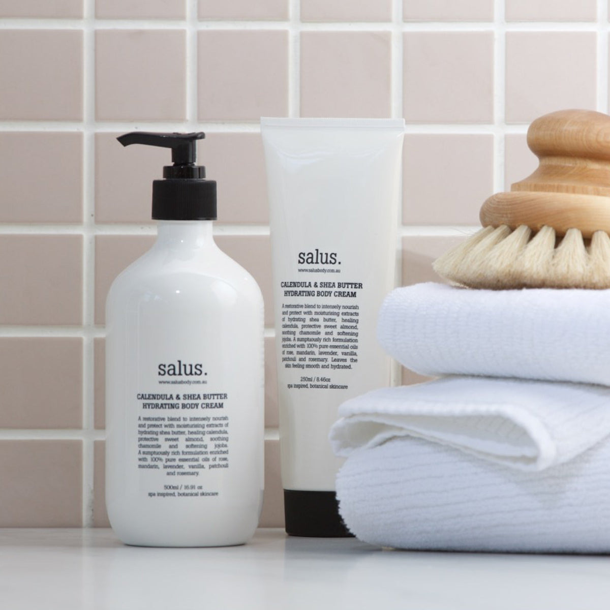 Salus Calendula & Shea Butter body cream pump bottle and tube on a tiled shelf with towels and a body brush