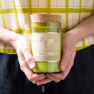 Person in a yellow check apron holding the green glass Cedar & Saffron candle in both hands
