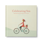 Cream hardcover titled Celebrating You with a woman cycling and a small dog in the basket