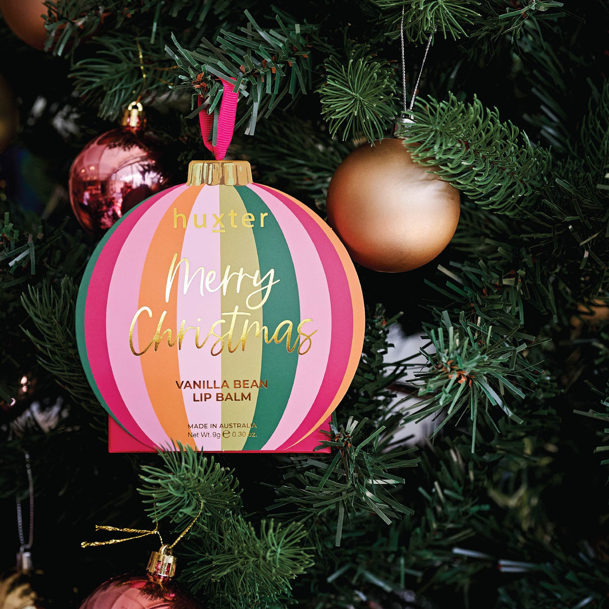 The Stripes bauble lip balm box hanging on a Christmas tree beside a gold bauble.