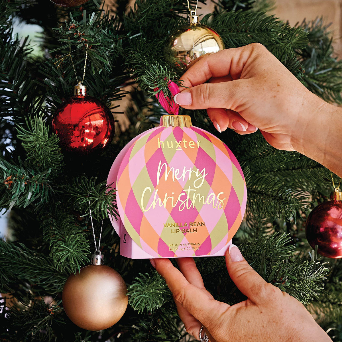 A hand hanging the Diamonds bauble lip balm box on a Christmas tree among red and gold baubles.